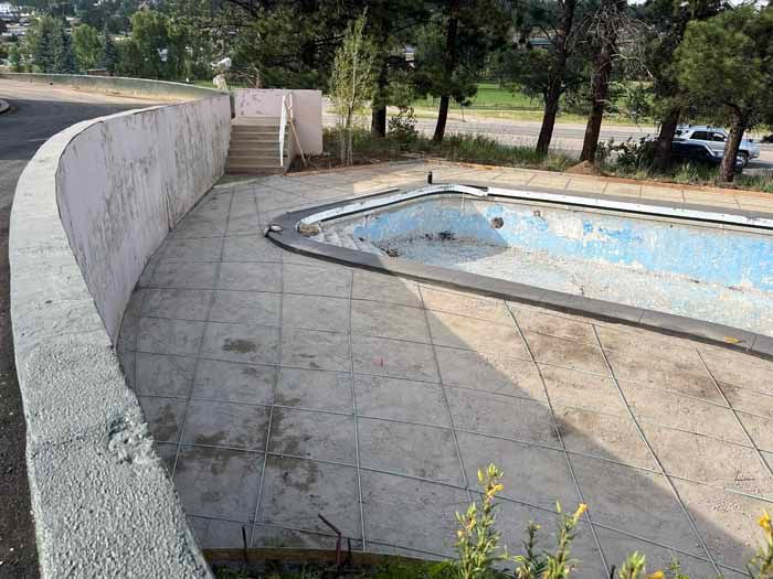 An empty, dilapidated swimming pool with a concrete surround and wall. Trees and hillside in the background.
