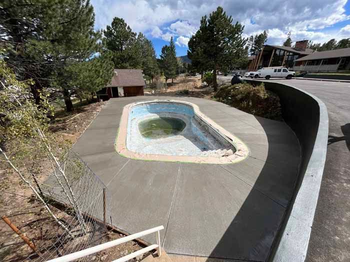 Empty, overgrown swimming pool surrounded by fresh concrete, trees, and a building on a sunny day.