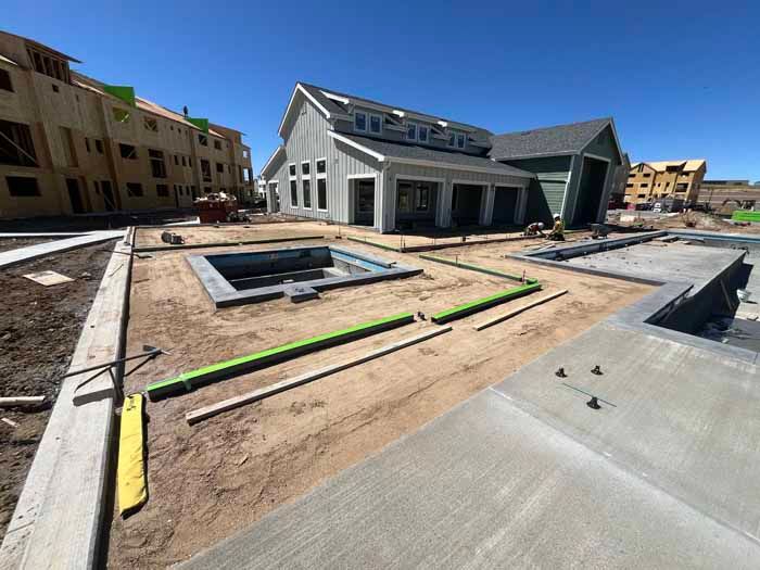 Construction site with a pool, building, and concrete.  Green pipes and dirt visible. Sunny day.