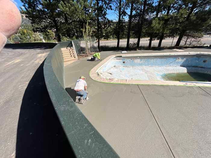 Two workers spread wet concrete around an empty swimming pool. Sunny outdoors.