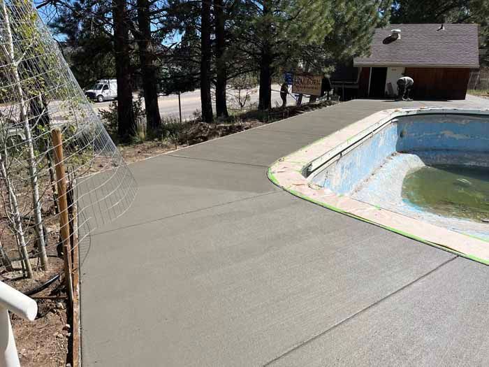 Newly poured concrete walkway next to a swimming pool, extending toward a small building.