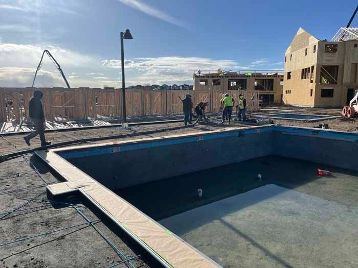Construction site: empty pool, workers, building frame, and concrete pump under a sunny sky.