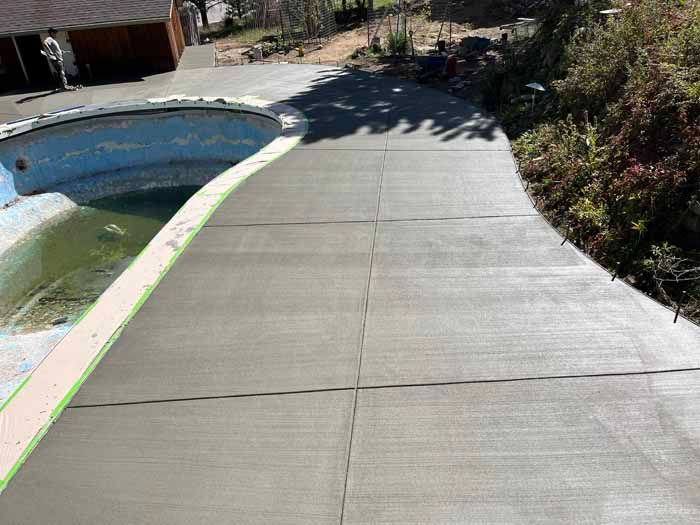 Newly poured concrete pathway next to an empty swimming pool. Construction worker in the background.