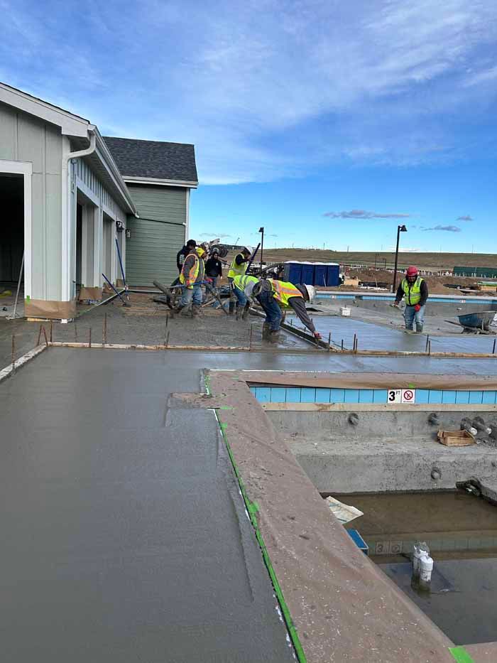 Construction workers pouring concrete around a pool, near a building with a blue sky.