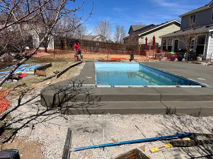 Pool construction: concrete pool with surrounding patio; workers in background, blue sky.