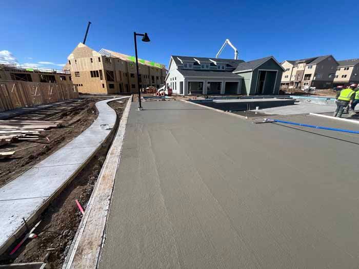 Construction site with freshly poured concrete road and sidewalk, buildings in background.