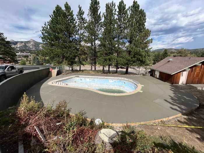 Newly poured concrete surrounding an empty, oval-shaped swimming pool. Tall trees stand behind the pool area.