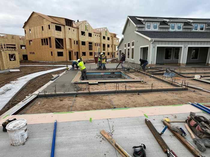 Construction site with workers around a pool; wooden buildings in the background and a cloudy sky.