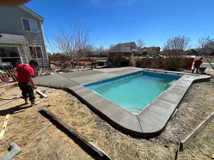 Workers build a concrete patio around a rectangular pool filled with turquoise water.