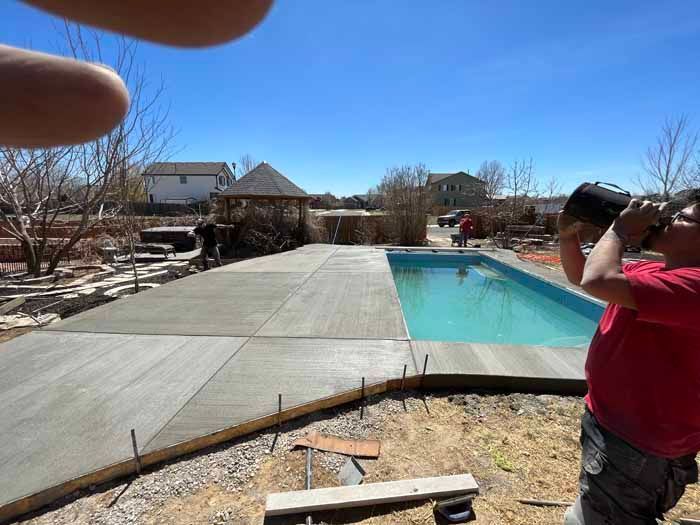 Construction worker drinks from a water jug by a newly constructed pool with concrete patio on a sunny day.