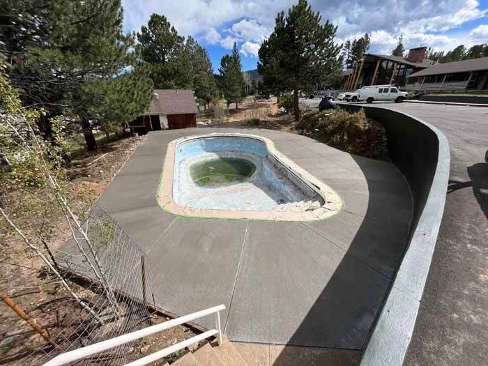 An empty, oval-shaped pool with new concrete surrounding it, set in a wooded area.