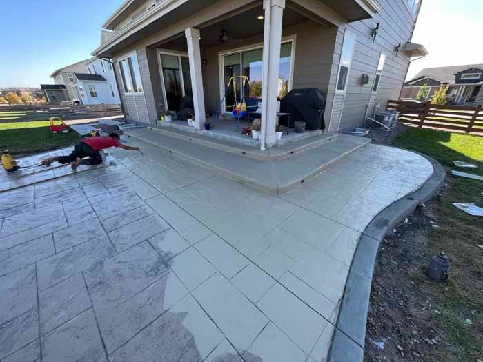 Construction worker applying finish to a new concrete patio and steps. Beige and gray colors, house in the background.