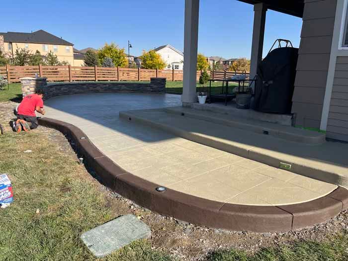 A worker kneels on a concrete patio. The patio has a brown border and is connected to a covered porch.