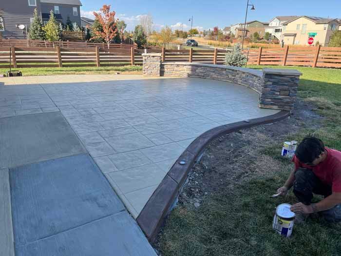 Man painting concrete border of a stamped patio with pillars and curved retaining wall in a yard.
