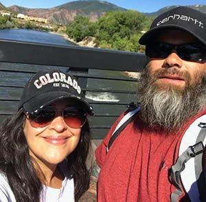Couple wearing hats and sunglasses, smiling for selfie outdoors. Mountains and water in the background.