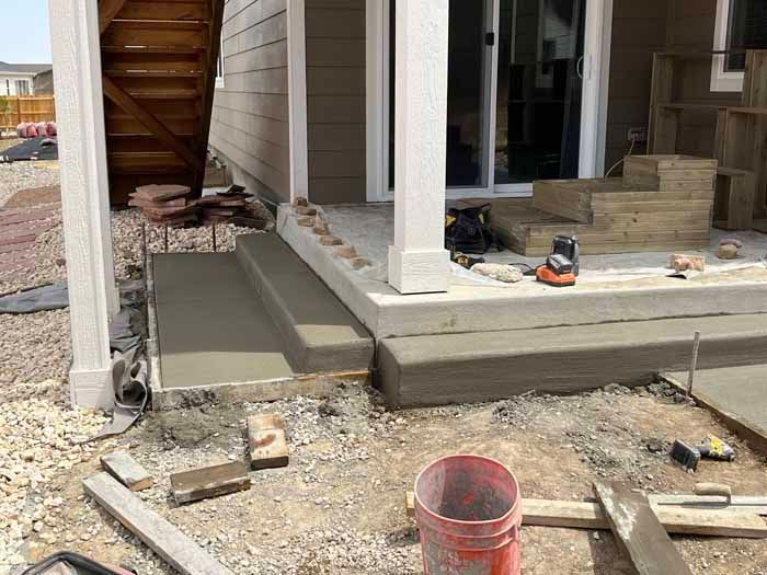 Concrete steps under construction outside a building with a white column, construction tools, and a wooden stairwell.