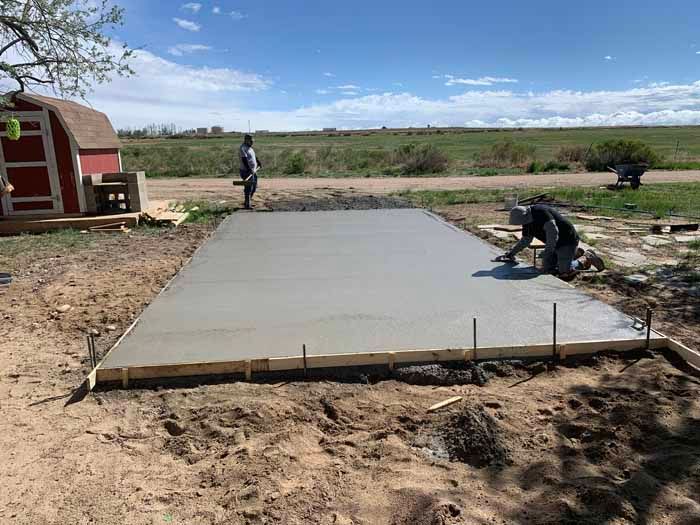 Workers finishing a concrete slab outdoors. One kneels smoothing the surface, another stands nearby, and a small red shed is visible.