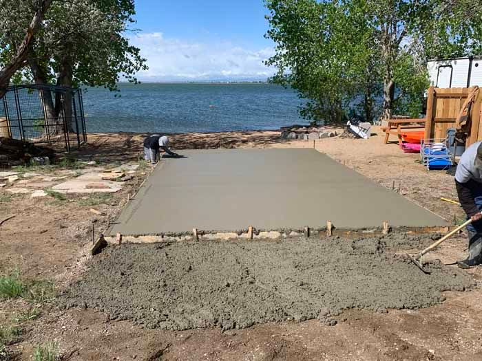 Workers pouring and smoothing fresh concrete slab near a body of water.