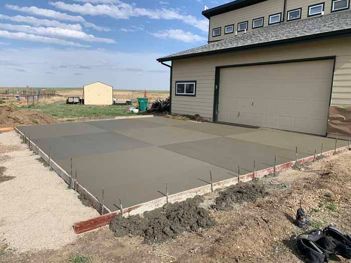 Freshly poured concrete slab next to a building with a garage door and small outbuilding in a field.