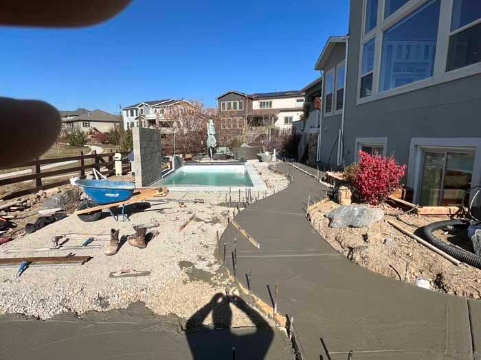 Backyard under construction with a newly poured concrete walkway next to a pool.