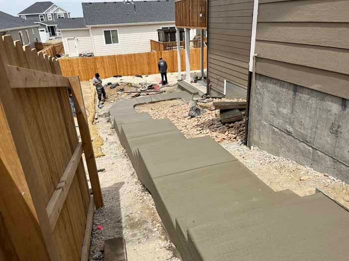 Workers constructing a concrete walkway between a wooden fence and a house.