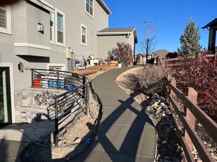 Concrete walkway beside a house with a fence and landscaping under a blue sky.