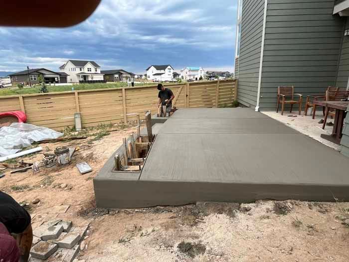 Workers smoothing a freshly poured concrete patio next to a house, with a wooden fence and houses in the background.