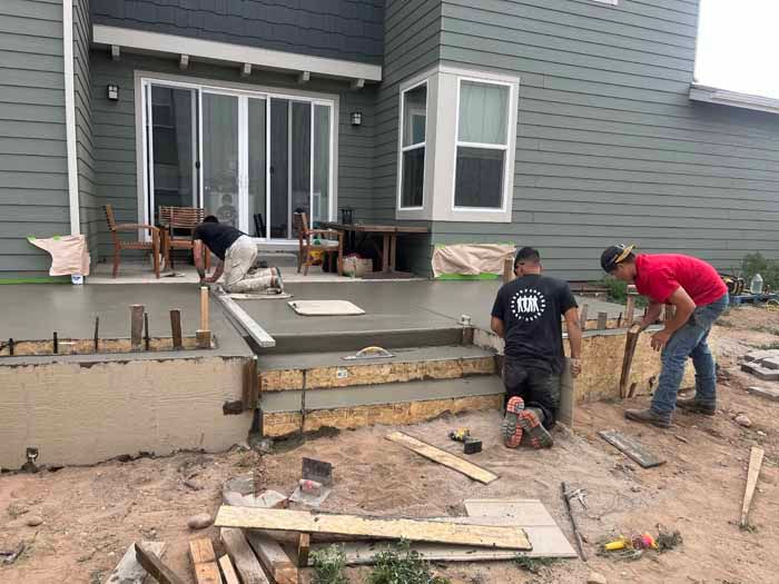Construction workers pouring concrete for steps on a house patio. They are kneeling, working in a dirt yard.
