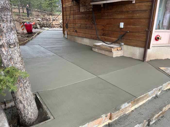 Newly poured concrete patio next to a wooden building, tree trunk in the corner.