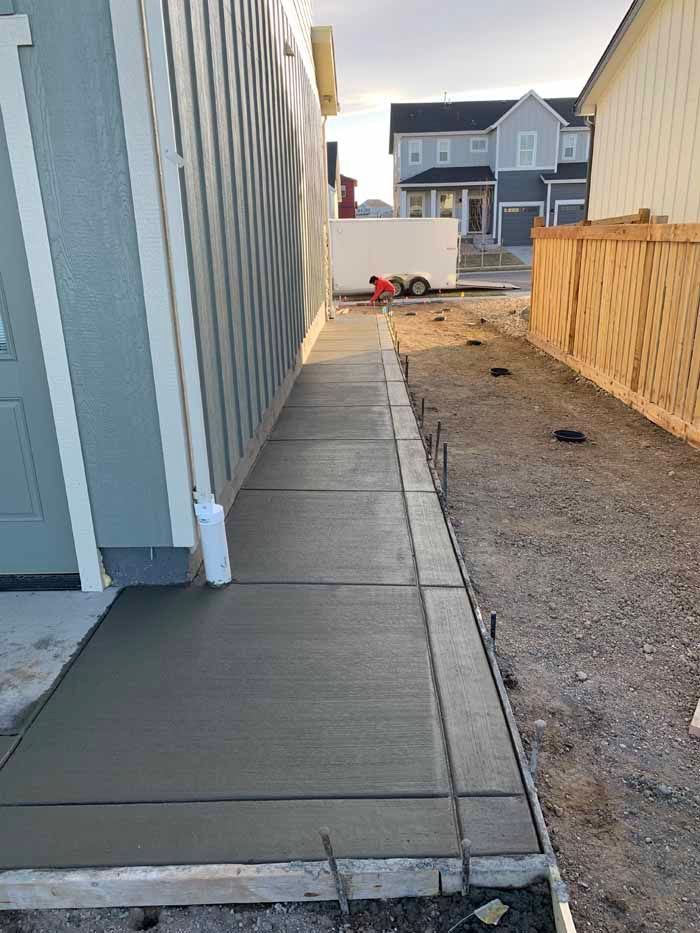 Newly poured concrete walkway next to a light blue building, with unfinished edges and wooden forms.