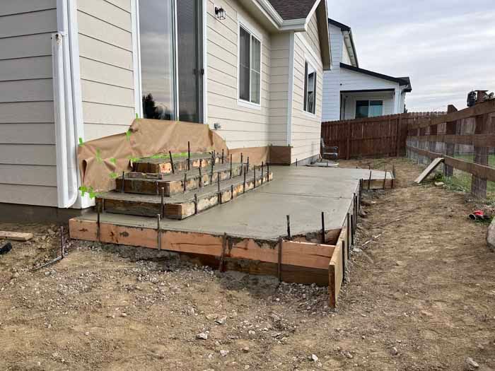 Concrete patio under construction with stairs and wooden forms, next to a house and fence.