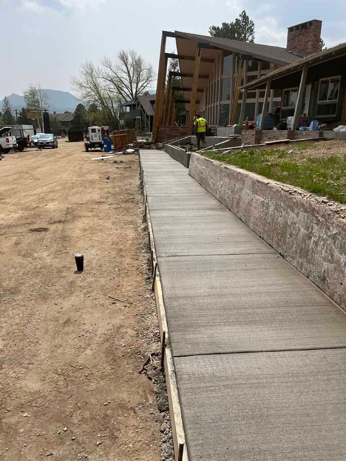 Newly poured concrete sidewalk with retaining wall next to dirt road, construction in the background.