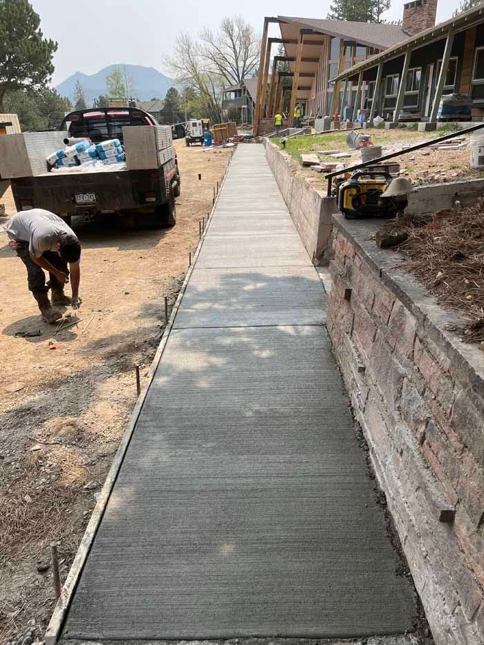 Sidewalk construction; worker smoothing wet concrete. Gray cement, dirt, truck, building, trees.
