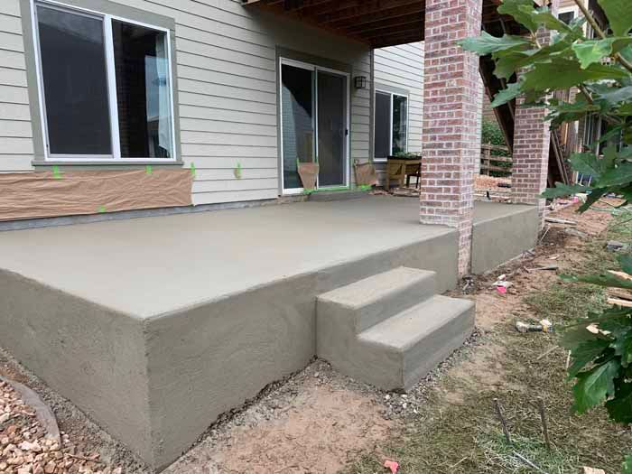 Newly poured concrete patio with steps leading up to a sliding glass door. A brick column supports a covered area.