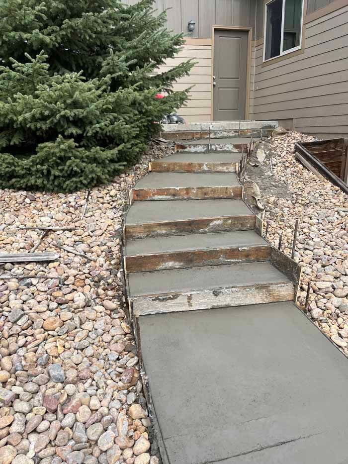 Concrete steps and walkway leading to a house entrance, bordered by gravel and a bush.