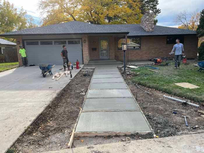 Workers pouring concrete sidewalk at a brick house, driveway on the left.