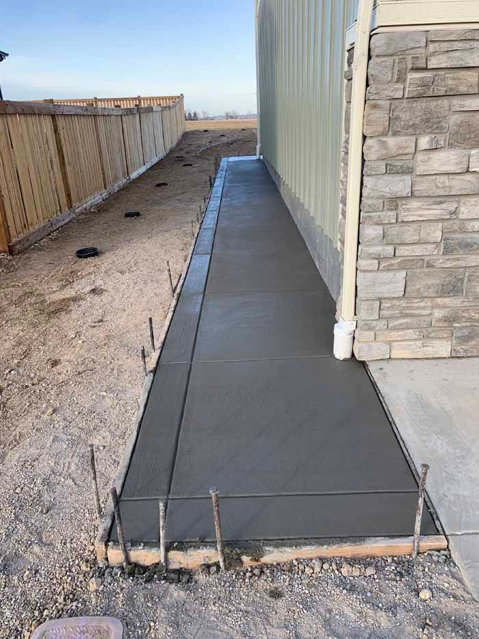 Freshly poured concrete walkway next to a building, with wooden forms and rebar visible.