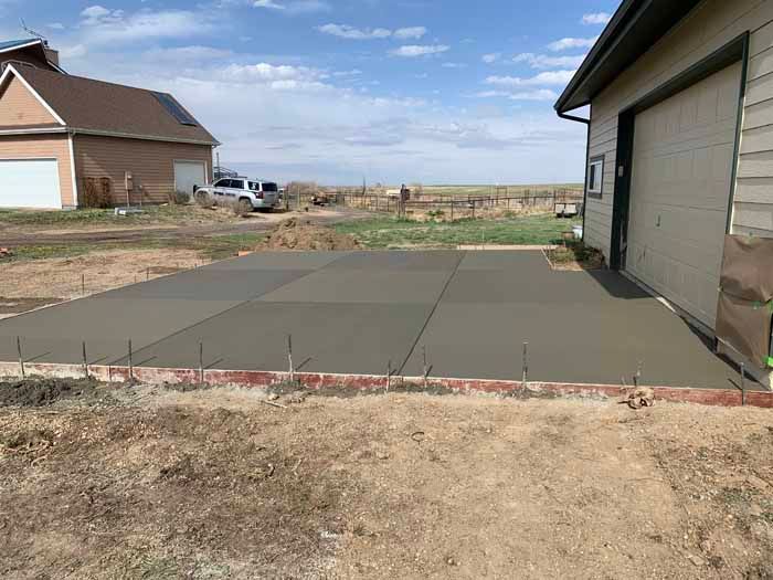 Newly poured concrete slab next to a garage, bordered by red bricks on dry, brown earth under a blue sky.