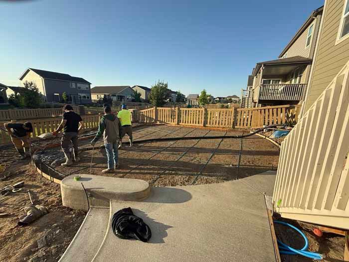 Construction workers in a backyard, pouring concrete. Wooden fence, houses, and blue sky.
