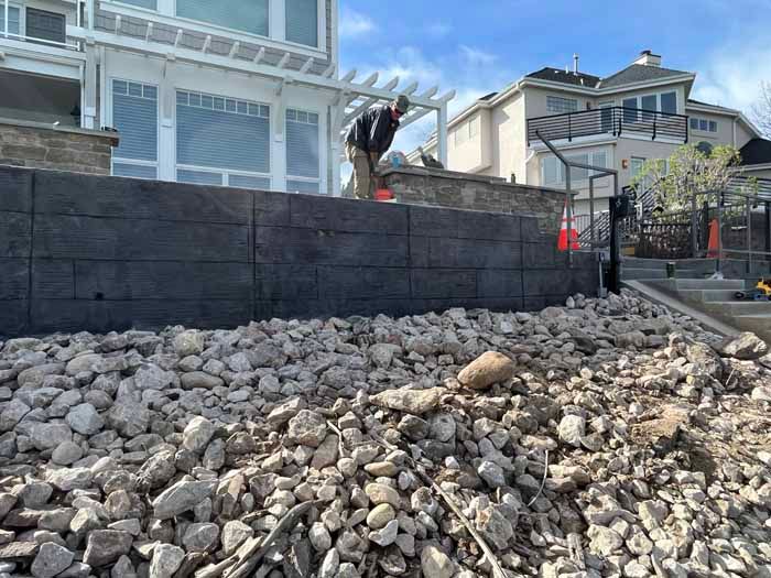 Construction worker on a retaining wall made of dark blocks, surrounded by gravel. Houses in background.