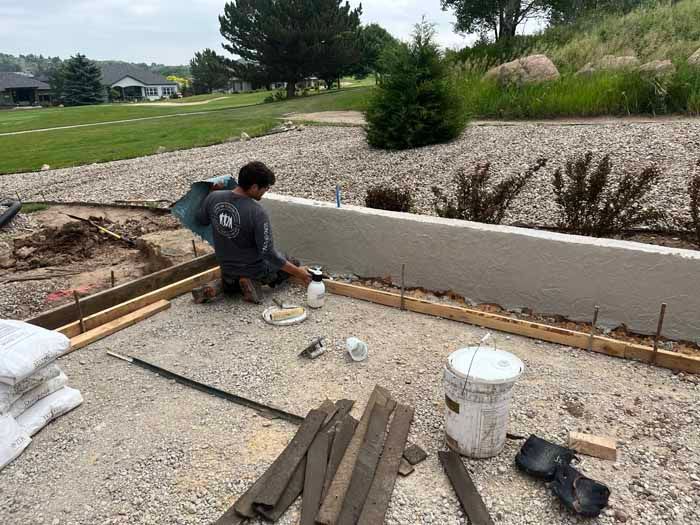 Construction worker applying sealant to concrete patio, near a retaining wall and grass.