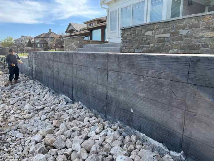 Concrete retaining wall next to rocks, partially obscuring a house with a person walking.