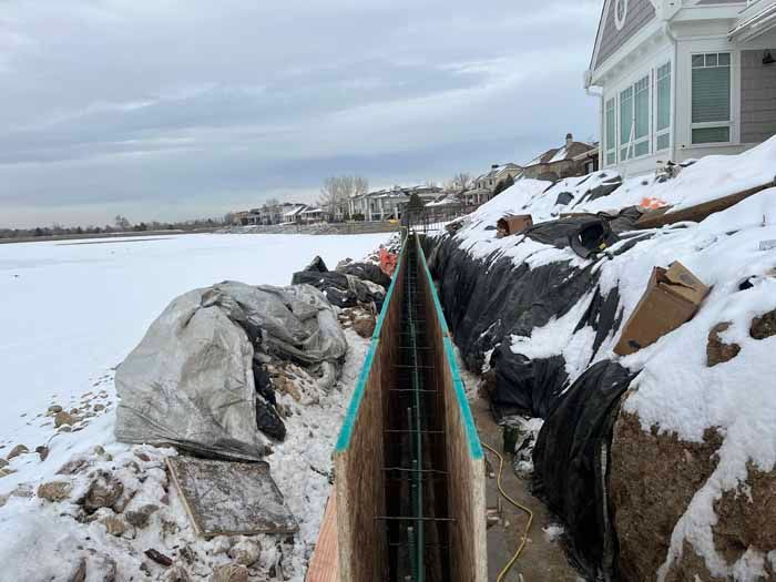 Trench dug along snow-covered shoreline, prepped for concrete. A house is visible in the background.
