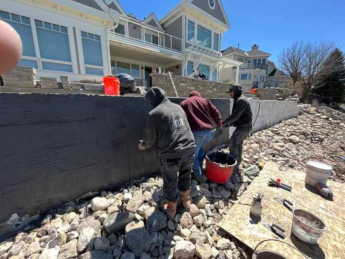 Construction workers applying material to a retaining wall near a lake, under sunny skies.