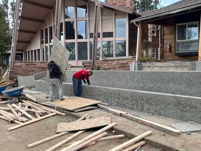 Construction workers building concrete steps near two buildings. One carries a large board, the other works on the steps.