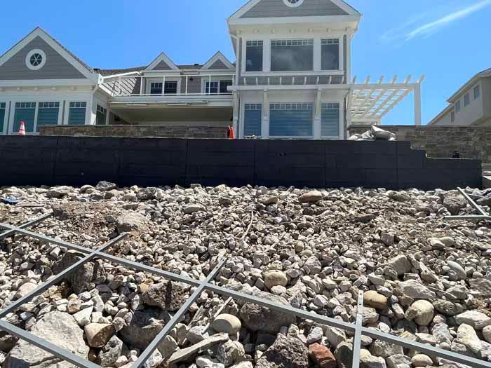 Construction site with large house in background, rubble and rebar in foreground, retaining wall being built.