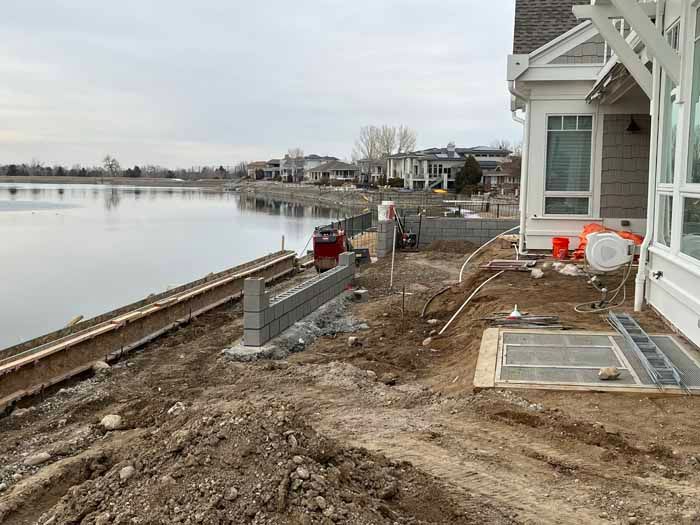Construction site near a lake. Concrete blocks form a wall, with dirt and equipment present. A white house is on the right.