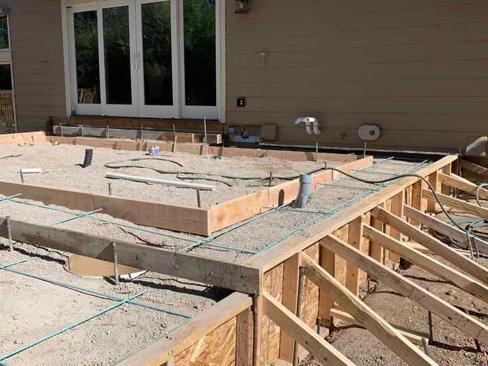 Construction site: wooden forms and rebar frame a concrete patio base next to a building.