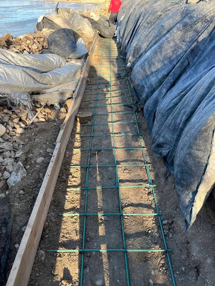 Construction site: Green rebar grid in trench, wooden form on the side, covered in black tarp.