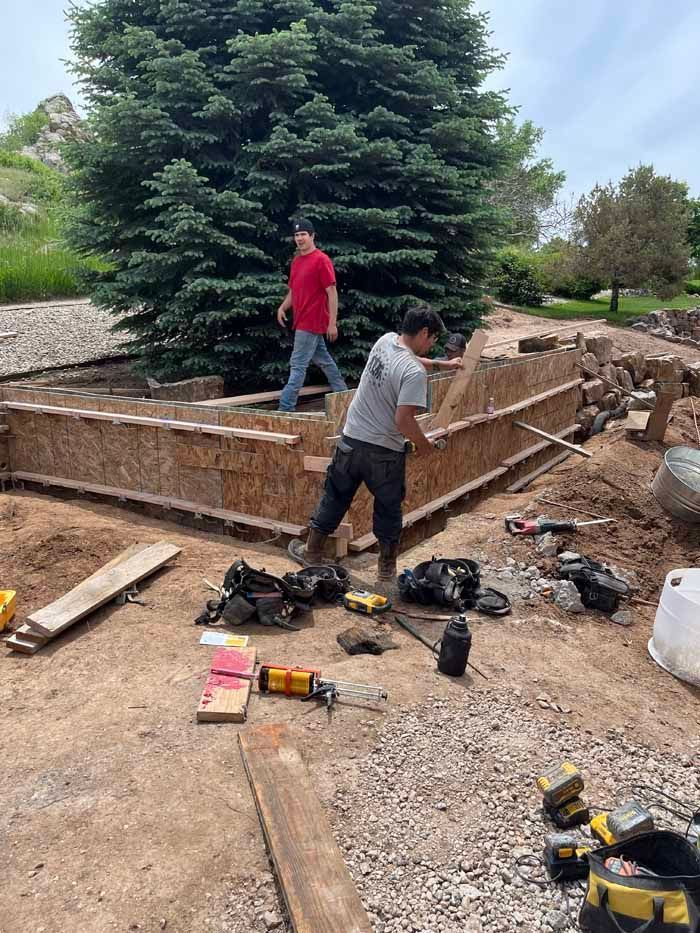 Two men constructing a wooden structure outdoors, surrounded by tools and dirt; a large tree in the background.
